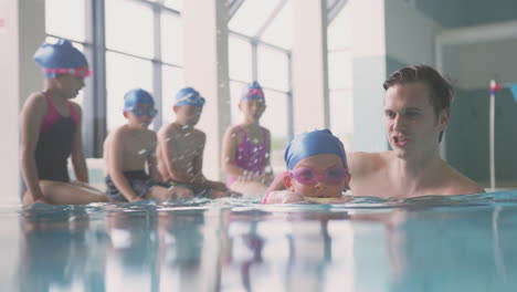 Male-Swimming-Coach-Giving-Girl-Holding-Float-Lesson-In-Pool