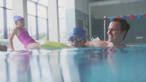 Male-Swimming-Coach-Giving-Boy-Holding-Float-Lesson-In-Pool