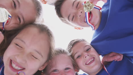 Low-Angle-View-Looking-Up-Into-Faces-Of-Children-In-Huddle-On-Sports-Day-Holding-Medals