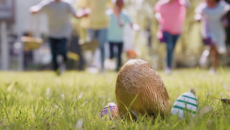 Grupo-De-Niños-Con-Orejas-De-Conejo-Corriendo-Para-Recoger-Huevos-De-Chocolate-En-La-Búsqueda-De-Huevos-De-Pascua-En-El-Jardín