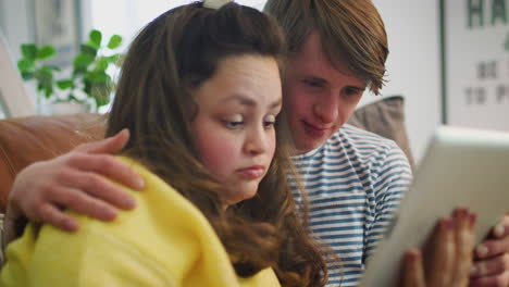 Junges-Paar-Mit-Down-Syndrom-Sitzt-Auf-Dem-Sofa-Und-Schaut-Zu-Hause-Auf-Ein-Digitales-Tablet