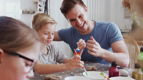 Parents-With-Children-Sitting-At-Table-Decorating-Eggs-For-Easter-At-Home