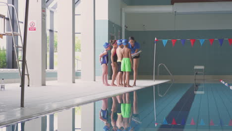 Female-Coach-Giving-Children-In-Swimming-Class-Briefing-As-They-Stand-On-Edge-Of-Indoor-Pool