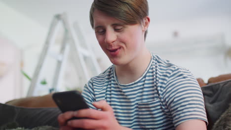 Young-Downs-Syndrome-Man-Sitting-On-Sofa-Using-Mobile-Phone-At-Home