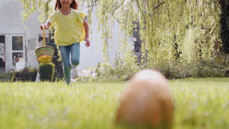 Girl-Wearing-Bunny-Ears-Running-To-Pick-Up-Chocolate-Egg-On-Easter-Egg-Hunt-In-Garden