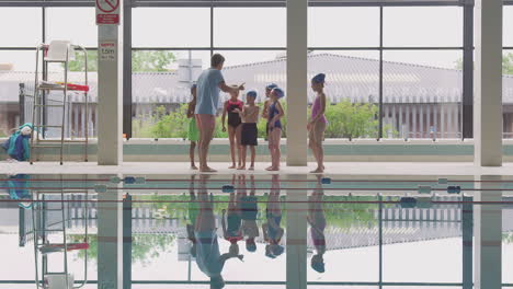 Male-Coach-Giving-Children-In-Swimming-Class-Instructions-As-They-Stand-On-Edge-Of-Indoor-Pool