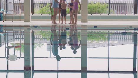 Male-Coach-Giving-Children-In-Swimming-Class-Instructions-As-They-Stand-On-Edge-Of-Indoor-Pool
