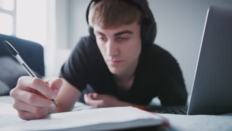 Male-College-Student-Wearing-Headphones-Works-On-Bed-In-Shared-House-With-Laptop