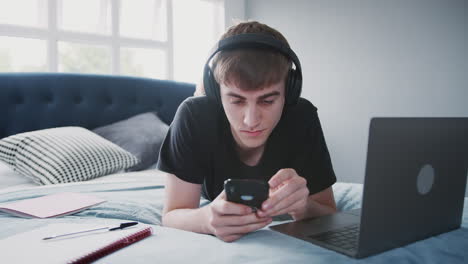 Male-College-Student-Wearing-Headphones-Works-On-Bed-In-Shared-House-With-Laptop-And-Mobile-Phone