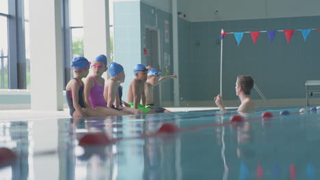Männlicher-Trainer-Im-Wasser-Gibt-Kindern-Anweisungen-Im-Schwimmunterricht,-Während-Sie-Am-Rand-Des-Hallenbades-Sitzen