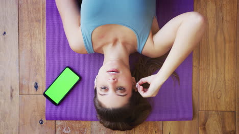 Overhead-View-Of-Woman-Lying-On-Exercise-Mat-At-Home-Taking-Out-Wireless-Earphones-After-Workout