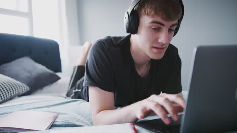 Male-College-Student-Wearing-Headphones-Works-On-Bed-In-Shared-House-With-Laptop