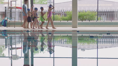 Male-Coach-Walks-Children-In-Swimming-Class-Along-Edge-Of-Indoor-Pool