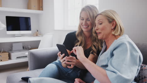Senior-Mother-With-Adult-Daughter-Sitting-On-Sofa-Looking-At-Pictures-On-Mobile-Phone