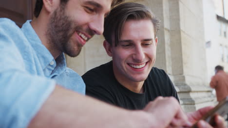 Male-Gay-Couple-Sitting-Outdoors-On-Steps-Of-Building-Looking-At-Mobile-Phones