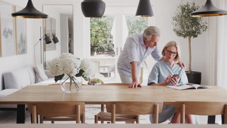 Senior-Couple-In-Dining-Room-At-Home-Looking-At-And-Mobile-Phone-Together