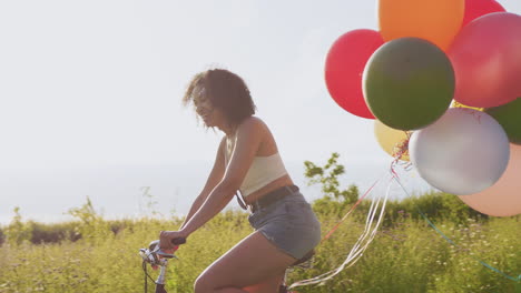 Junge-Frau-Fährt-Mit-Luftballons-Geschmücktes-Fahrrad-Durch-Die-Landschaft-Vor-Der-Gleißenden-Sonne