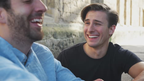 Male-Gay-Couple-Sitting-Outdoors-On-Steps-Of-Building-Laughing-Together
