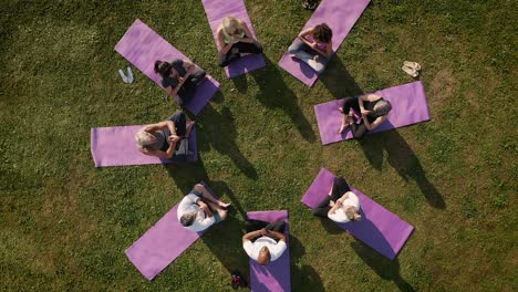 Drone-Shot-Of-Teacher-Leading-Group-Of-Mature-Men-And-Women-In-Class-At-Outdoor-Yoga-Retreat