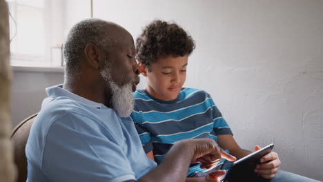Grandfather-Sitting-In-Chair-As-Grandson-Explains-How-To-Use-Digital-Tablet