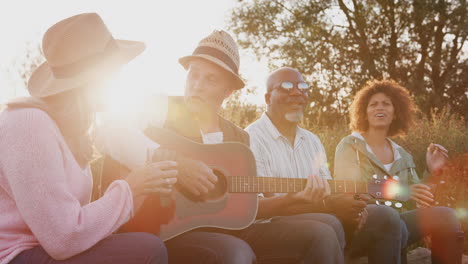 Group-Of-Mature-Friends-Sitting-Around-Fire-As-They-Drink-And-Sing-Songs-At-Outdoor-Campsite