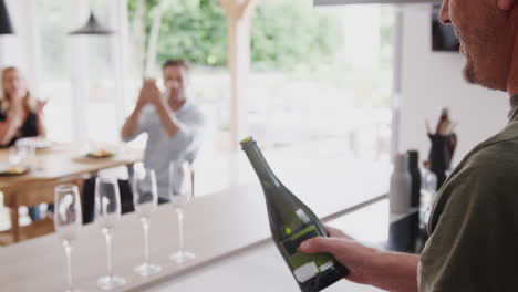 Senior-Man-Opens-Bottle-Of-Champagne-As-Family-With-Adult-Offspring-Eat-Meal-Around-Table-At-Home