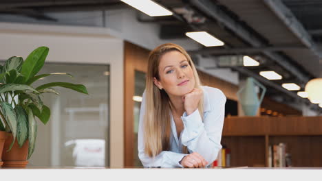 Portrait-Of-Smiling-Casually-Dressed-Young-Businesswoman-Standing-In-Modern-Open-Plan-Workplace
