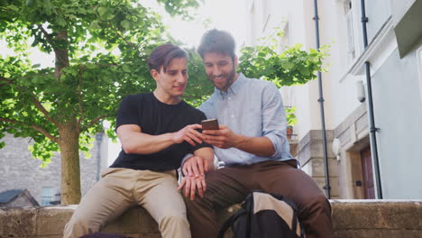 Male-Gay-Couple-Sitting-Outdoors-On-Wall-Of-Building-Looking-At-Mobile-Phone