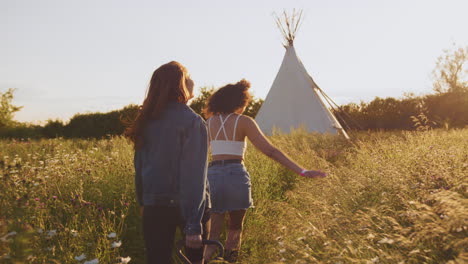 Two-Female-Friends-Walking-Pulling-Trolley-Through-Field-Towards-Teepee-On-Summer-Camping-Vacation