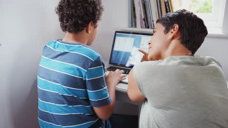 Rear-View-Of-Mother-Helping-Son-Using-Laptop-On-Desk-In-Bedroom