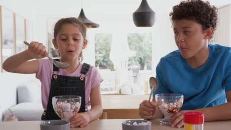 Children-In-Kitchen-At-Home-Eating-Ice-Cream-And-Pulling-Faces-At-Each-Other