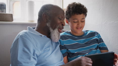 Grandfather-Sitting-In-Chair-As-Grandson-Explains-How-To-Use-Digital-Tablet