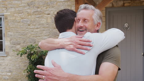 Senior-Father-Greets-And-Hugs-Adult-Son-Outside-Front-Door-Of-House-As-He-Visits
