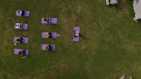 Drone-Shot-Of-Teacher-Leading-Group-Of-Mature-Men-And-Women-In-Class-At-Outdoor-Yoga-Retreat