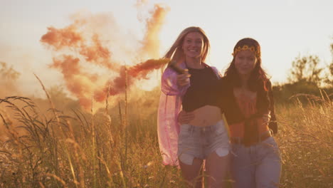 Two-Female-Friends-Camping-At-Music-Festival-Running-Through-Field-With-Smoke-Flares