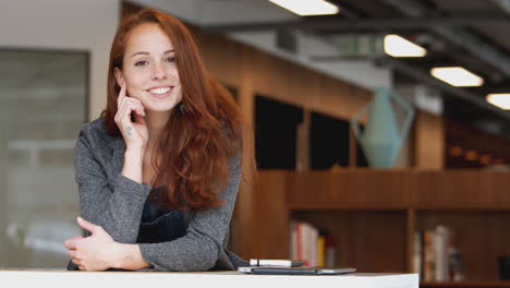 Portrait-Of-Casually-Dressed-Young-Businesswoman-Standing-In-Modern-Open-Plan-Workplace
