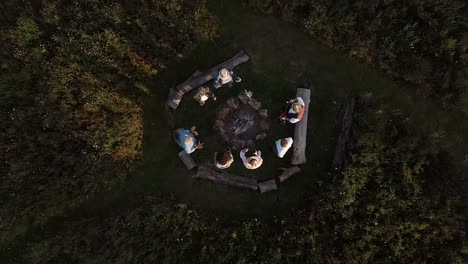 Drone-Shot-Of-Mature-Friends-Making-Toast-As-They-Sit-Around-Fire-At-Outdoor-Campsite