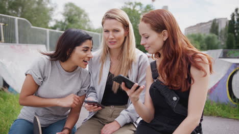 Three-Female-Friends-Looking-At-Mobile-Phone-In-Urban-Skate-Park-And-Laughing