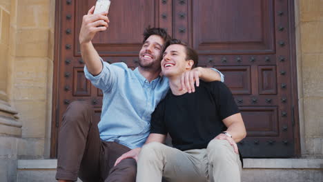 Una-Pareja-Gay-Masculina-Tomando-Selfie-En-Un-Teléfono-Móvil-Sentado-Al-Aire-Libre-En-Las-Escaleras-Del-Edificio