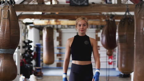 Portrait-Of-Female-Boxer-In-Gym-Walking-Towards-Camera-Past-Old-Fashioned-Punching-Bags