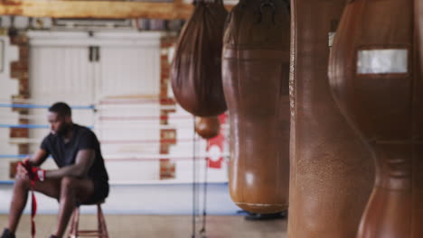 Männlicher-Boxer-Trainiert-Im-Fitnessstudio-Und-Legt-Wraps-Auf-Die-Hände,-Die-Neben-Einem-Boxring-Und-Boxsäcken-Sitzen