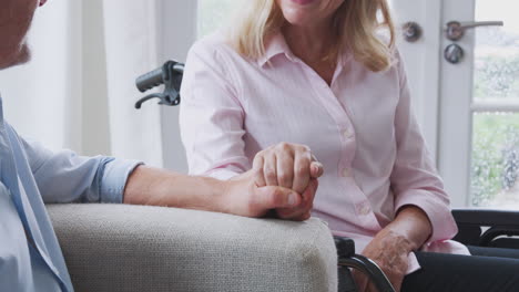 Close-Up-Of-Senior-Couple-With-Woman-In-Wheelchair-Sitting-In-Lounge-At-Home-Talking-Together