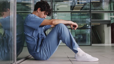 Tired-Young-Male-Doctor-Wearing-Scrubs-Sitting-Against-Wall-In-Modern-Hospital-Building