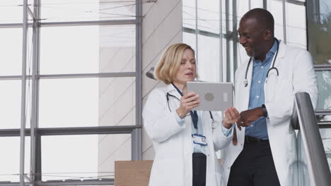 Male-And-Female-Doctors-Having-Informal-Meeting-In-Modern-Hospital-Looking-At-Digital-Tablet