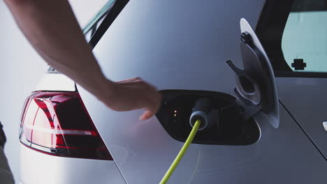 Close-Up-Of-Hand-Attaching-Power-Cable-To-Electric-Car-Before-Taking-Cloth-Bags-From-Trunk