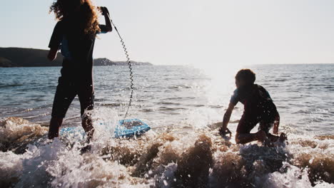Two-Silhouetted-Children-Wearing-Wetsuits-Playing-In-Sea-With-Bodyboards-On--Summer-Beach-Vacation