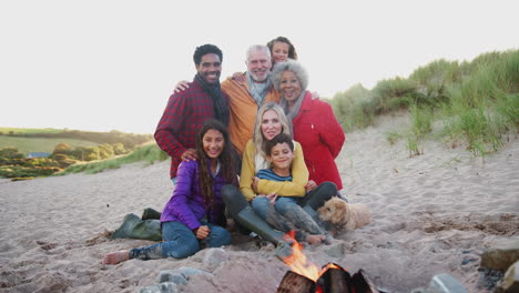 Portrait-Of-Active-Multi-Generation-Family-With-Pet-Dog-Sitting-By-Fire-On-Winter-Beach-Vacation