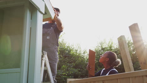 Male-Carpenter-With-Female-Apprentice-Putting-Roof-On-Outdoor-Summerhouse-In-Garden