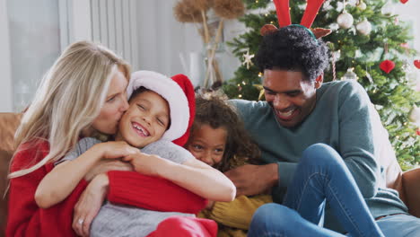 Parents-Tickling-Children-As-Family-Sit-On-Sofa-Celebrating-Christmas-Together