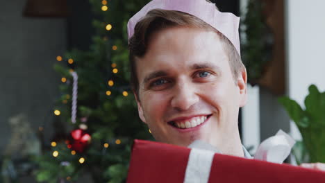 Porträt-Eines-Mannes-Mit-Papierhut,-Der-Einen-Stapel-Weihnachtsgeschenke-Trägt-Und-Zu-Hause-Am-Baum-Steht